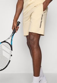 Ljusgula träningsshorts i mjukt tyg, med sidofickor och "Sergio Tacchini"-logotyp i svart. Håller en tennisracket.