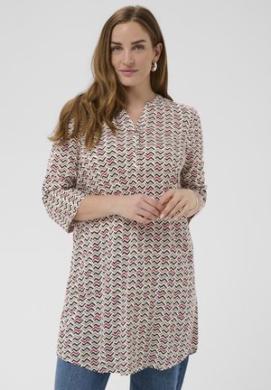 Femeie cu păr lung, castaniu, purtând cercei cerc și o tunică bej cu model, cu mâneci trei sferturi, și blugi albaștri, stând pe fundal gri.