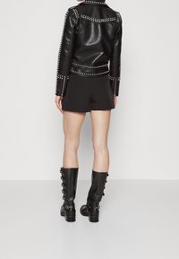 Veste en cuir noir avec des clous en métal argenté, associée à un short noir et des bottes montantes avec des accents de boutons. Texture lisse et design ajusté.