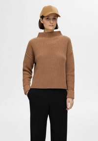 Brauner gestrickter Pullover mit hohem Kragen und langen Ärmeln, getragen mit schwarzen weit geschnittenen Hosen. Enthält eine passende braune Mütze.