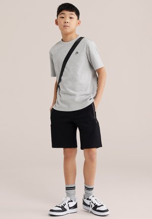 Băiat stând cu mâinile în buzunare, purtând un tricou gri, pantaloni scurți negri, șosete cu dungi, adidași alb-negru și o geantă transversală.