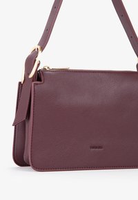 Borsa a tracolla in pelle bordeaux con finitura testurizzata. Presenta una tracolla removibile e accessori in tonalità oro. Chiusura con zip e dettaglio di branding impresso.
