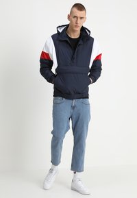 Marine let jakke med hvide og røde detaljer, foret hætte, elastiske manchetter og en frontlomme. Båret med lyseblå jeans og hvide sneakers.