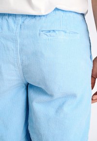 Pantalones de pana de color azul claro con estrechas rayas verticales, que cuentan con un bolsillo trasero y una textura suave, con una mano parcialmente visible en la cintura.