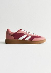 Sneaker rosa e marrone scuro con texture scamosciata, caratterizzata da tre righe bianche e una suola in gomma marrone. Design con lacci, silhouette bassa.