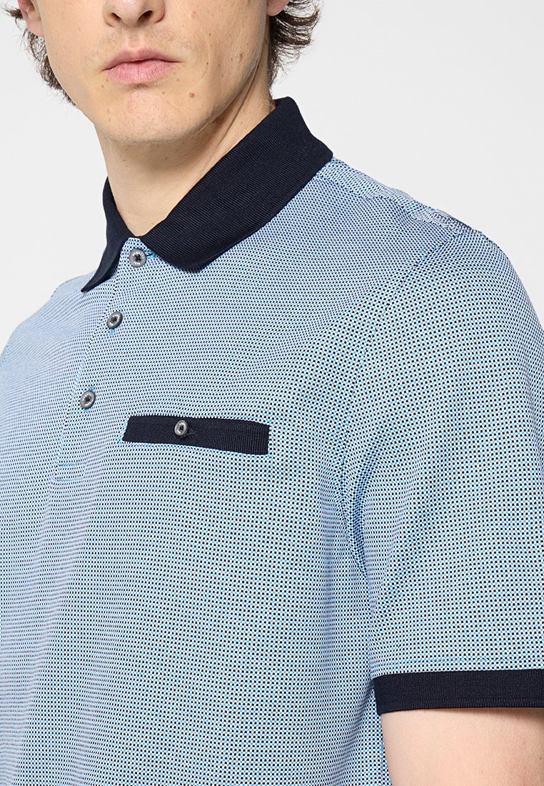 Polo bleu et blanc à motifs avec un col marine, trois boutons et une poche poitrine agrémentée d'un accent foncé.