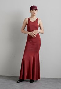 Červené priliehavé maxi šaty s rozšíreným lemom, vyrobené z hladkej látky. Skombinované s ladiacou čiapkou a čiernymi topánkami na neutrálnom pozadí.
