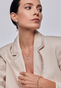 Złoty naszyjnik z przezroczystym kamieniem wisiorkiem, warstwowy złoty bransoletka i złoty pierścionek. Ubrany z beżową marynarką z widocznym przeszyciem.