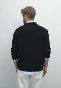 Schwarze Woll-Cardigan mit lässigem Schnitt, gerippten Bündchen und einem sauberen Finish. Über einem hellblauen gestreiften Hemd und weißen Hosen getragen.