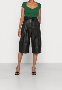 Zwarte leren culottes met een brede, losse pasvorm, voorzien van een ceintuur in de taille en plooien. Gepaard met een kort groen ribbeltopje en zwarte sandalen.