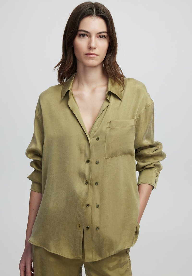 Camisa verde oliva de botones, hecha de un tejido ligero. Presenta un bolsillo en el pecho, mangas largas y un corte relajado con una textura suave.