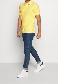 Keltainen polo-paita kauluksella ja kahdella napilla, yhdistettynä kapealahkeisiin tummansinisiin farkkuihin. Valkoiset tennarit tummansinisillä yksityiskohdilla viimeistelevät asun.