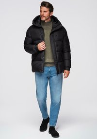 Chaqueta acolchada negra con capucha que cuenta con cierre de cremallera y botones a presión, llevada sobre un suéter de punto verde y jeans azules. Textura suave, diseño acolchado.