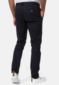 Chinos navy con pierna entallada, que cuentan con un bolsillo trasero con cierre de botón y una etiqueta de cuero marrón claro en la cinturilla. Llevado con zapatos blancos.