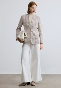 Veste en tweed texturée rose et crème avec un revers cranté, un bouton unique, associée à un pantalon blanc à jambes larges et un sac crème.
