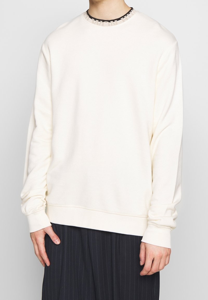 Felpa leggera beige chiaro con collo a giro e polsini a costine. Il tessuto sembra morbido e presenta una vestibilità comoda. Abbinata a pantaloni a righe.