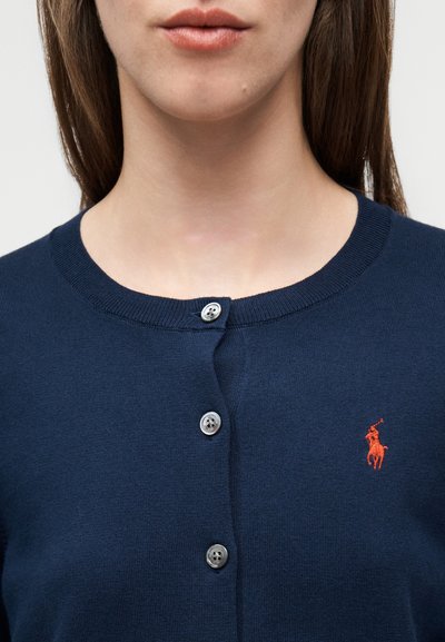 Καρντιγκάν σε ναυτικό μπλε χρώμα από πλεκτό υλικό. Διαθέτει στρογγυλή λαιμόκοψη, τέσσερα ασημένια κουμπιά και κεντημένο πορτοκαλί λογότυπο στο στήθος.