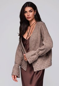 Braune gestrickte Strickjacke mit weit ausgestellten Ärmeln, getragen über einem braunen Satinkleid. Strukturierter Stoff und lockere Passform erzeugen einen geschichteten Look.