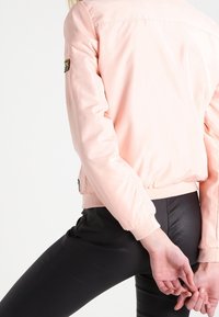 Jasnoróżowa kurtka bomberka wykonana z gładkiego materiału, z prążkowanym wykończeniem u dołu i na mankietach, z naszywką na lewym rękawie.