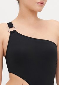 Maillot de bain noir asymétrique en tissu lisse, avec un détail en anneau argenté à l'épaule et un design de jambe haute.