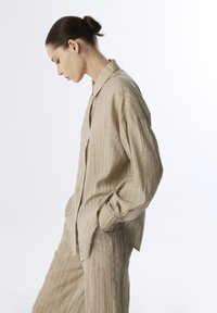 Beige linnen shirt met een ontspannen pasvorm, voorzien van verticale strepen en een subtiele textuur. Model poseert zijwaarts, met handen in de zakken.
