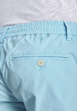 Hellblaue elastische Bundhose mit einer Gesäßtasche, die durch einen weißen Knopf gesichert ist, getragen zu einem in die Hose gesteckten weißen Hemd.