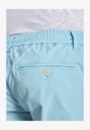 Hellblaue elastische Bundhose mit einer Gesäßtasche, die durch einen weißen Knopf gesichert ist, getragen zu einem in die Hose gesteckten weißen Hemd.