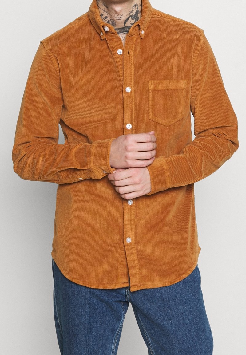 Camicia in corda marrone con colletto a bottone, dotata di tasca sul petto e bottoni bianchi, indossata sopra jeans blu. Tessuto testurizzato con righe verticali.