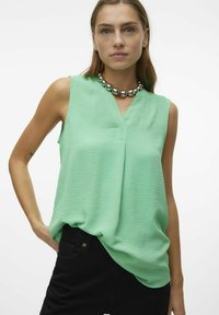 Blouse sans manches vert menthe avec un décolleté en V, fabriquée à partir d'un tissu texturé. Portée avec un collier métallique volumineux et un jean noir.