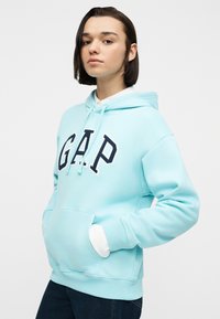 Νεαρός με κοντά σκούρα μαλλιά που φοράει ένα ανοιχτό μπλε φούτερ GAP και σκούρο τζιν, στέκεται με τα χέρια στις τσέπες μπροστά σε απλό φόντο.