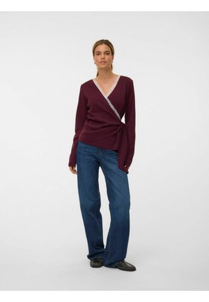 Kvinde står iført en langærmet bordeaux wrap-top med blondekanter, blå jeans og sorte sko mod en ensfarvet hvid baggrund.