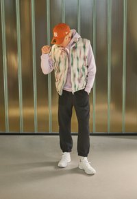 Gepolsterte Weste mit grünen und pinken Streifen über einem hellvioletten Hoodie, kombiniert mit schwarzen Hosen und weißen Sportschuhen. Eine orange Kappe rundet das Outfit ab.