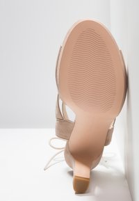 Chaussure à talon haut en suède nude avec un embout allongé, un talon fin et une semelle texturée. Elle présente une lanière avec des lacets autour de la cheville.