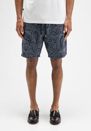 Mann trägt marineblaue Shorts mit schwarzen abstrakten Linienmustern, weißes T-Shirt und dunkelbraune Lederloafers, steht vor weißem Hintergrund.