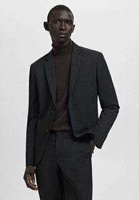 Man draagt een donker colbert, bijpassende broek en een donkere coltrui, staand tegen een effen lichte achtergrond.