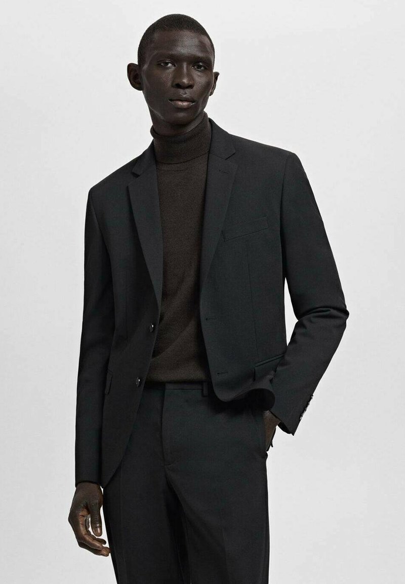 Man draagt een donker colbert, bijpassende broek en een donkere coltrui, staand tegen een effen lichte achtergrond.