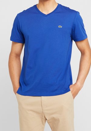 T-shirt en coton bleu avec un col en V et des manches courtes. Présente un petit logo vert sur la poitrine, une texture lisse et une coupe décontractée.