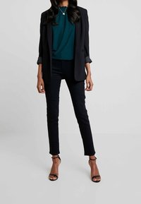 Svart kavaj över en teal texturerad blus, tillsammans med svarta slim-fit byxor och svarta sandaler med remmar. En enkel silverhalskedja lägger till detaljer.