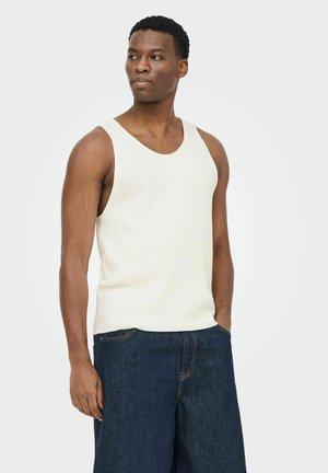 Joven con cabello corto, vestido con una camiseta blanca sin mangas y jeans azul oscuro, de pie con una mano en el bolsillo contra un fondo claro.