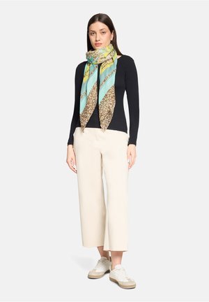 Donna in piedi che indossa una maglia nera a maniche lunghe, pantaloni beige a gamba larga, sneakers bianche e una sciarpa avvolta intorno al collo con motivi blu, gialli e maculati.