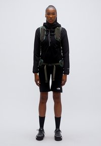 Μαύρο υπαίθριο μπουφάν με κουκούλα, πράσινα λουριά σακιδίου, μαύρο σορτς και μαύρες κάλτσες. Φέρει το λογότυπο The North Face στο σορτς και τα παπούτσια.