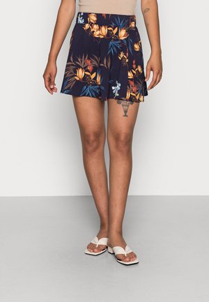 Personne portant un short à fleurs bleu marine et des sandales beiges ouvertes, montrant un tatouage sur la cuisse gauche.