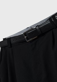 Ceinture en cuir noir avec boucle argentée passée dans les passants d'un pantalon noir avec une ceinture intérieure rayée.