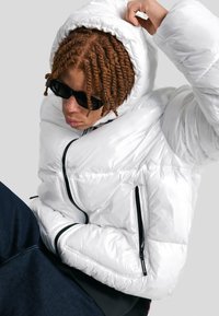 Persoană tânără cu părul împletit, purtând ochelari de soare negri și o jachetă puf din material lucios alb, cu glugă, ajustând gluga în timp ce este așezată.