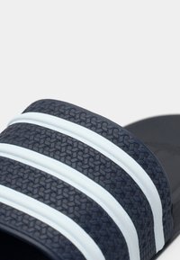 Șlapi de culoare albastru închis cu dungi albe, partea superioară din cauciuc texturat și talpă netedă. Prezintă un model geometric pentru aderență.