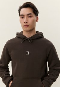 Giovane uomo con capelli corti e scuri che indossa una felpa con cappuccio marrone scuro con un logo bianco "II", in piedi davanti a uno sfondo beige uniforme.