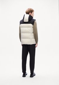 Puffer vest i kremfarge og svart, med hette, quiltet design og myk tekstur. Båret over en langermet skjorte og kombinert med mørke bukser.