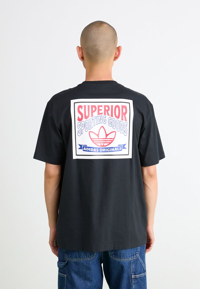 T-shirt en coton noir avec un grand motif au dos affichant le texte "SUPERIOR SPORTING GOODS" et le logo Adidas, coupe décontractée.