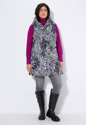 Gilet à motif noir et blanc avec un grand col superposé sur un col roulé violet, associé à des leggings gris et des bottes noires jusqu'au genou.