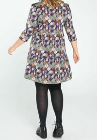 Robe multicolore avec un motif géométrique de feuilles, dotée de manches trois-quarts et d'une jupe évasée. Portée avec des collants et des chaussures noirs.